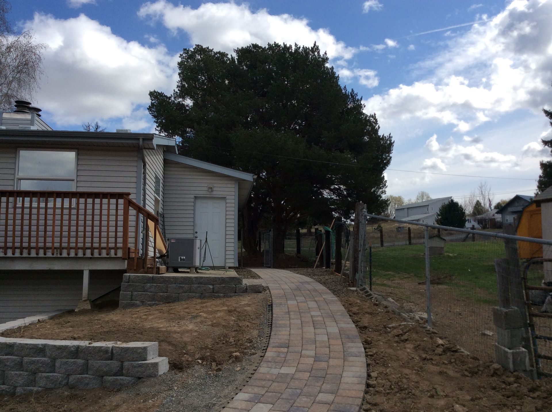 A paved stone walkway leads toward a light-colored house with a wooden deck, situated next to a large evergreen tree.