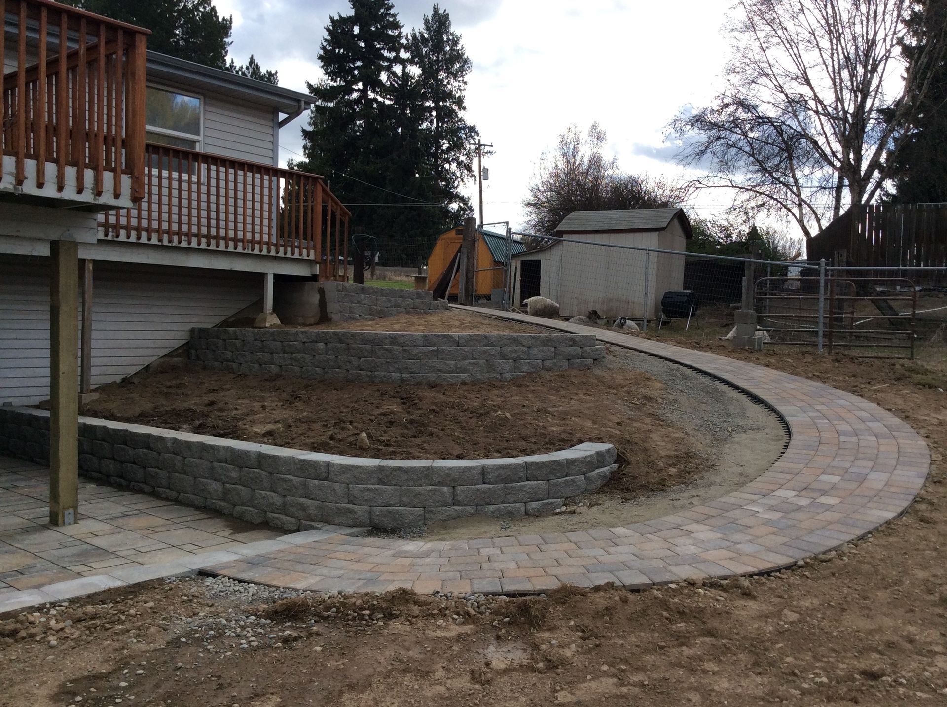 A two-tier stone retaining wall stands in a backyard next to a house with a wooden deck and a curved paved walkway.
