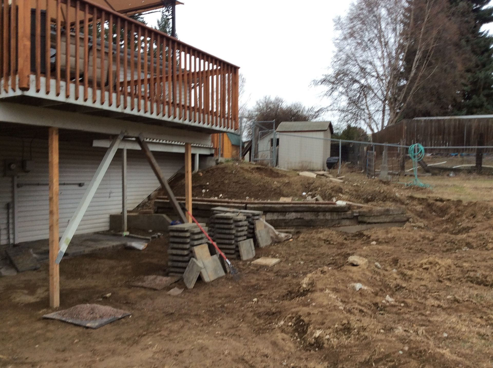 A wooden deck stands above a dirt yard under construction, featuring stacked paving stones and a low retaining wall.
