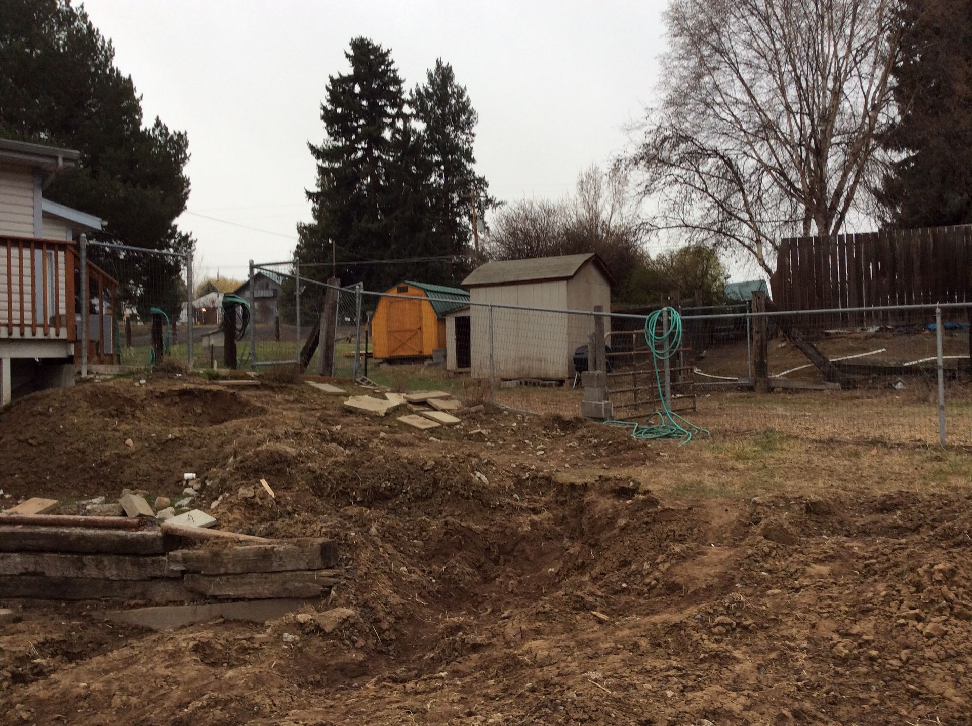 A backyard construction site with a freshly dug trench, a shed, a yellow tarp, and a wooden fence under a cloudy sky.
