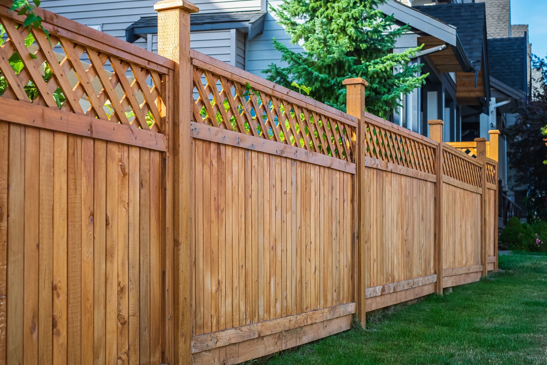 A wooden privacy fence with a lattice top section runs along a grassy lawn in front of a house.