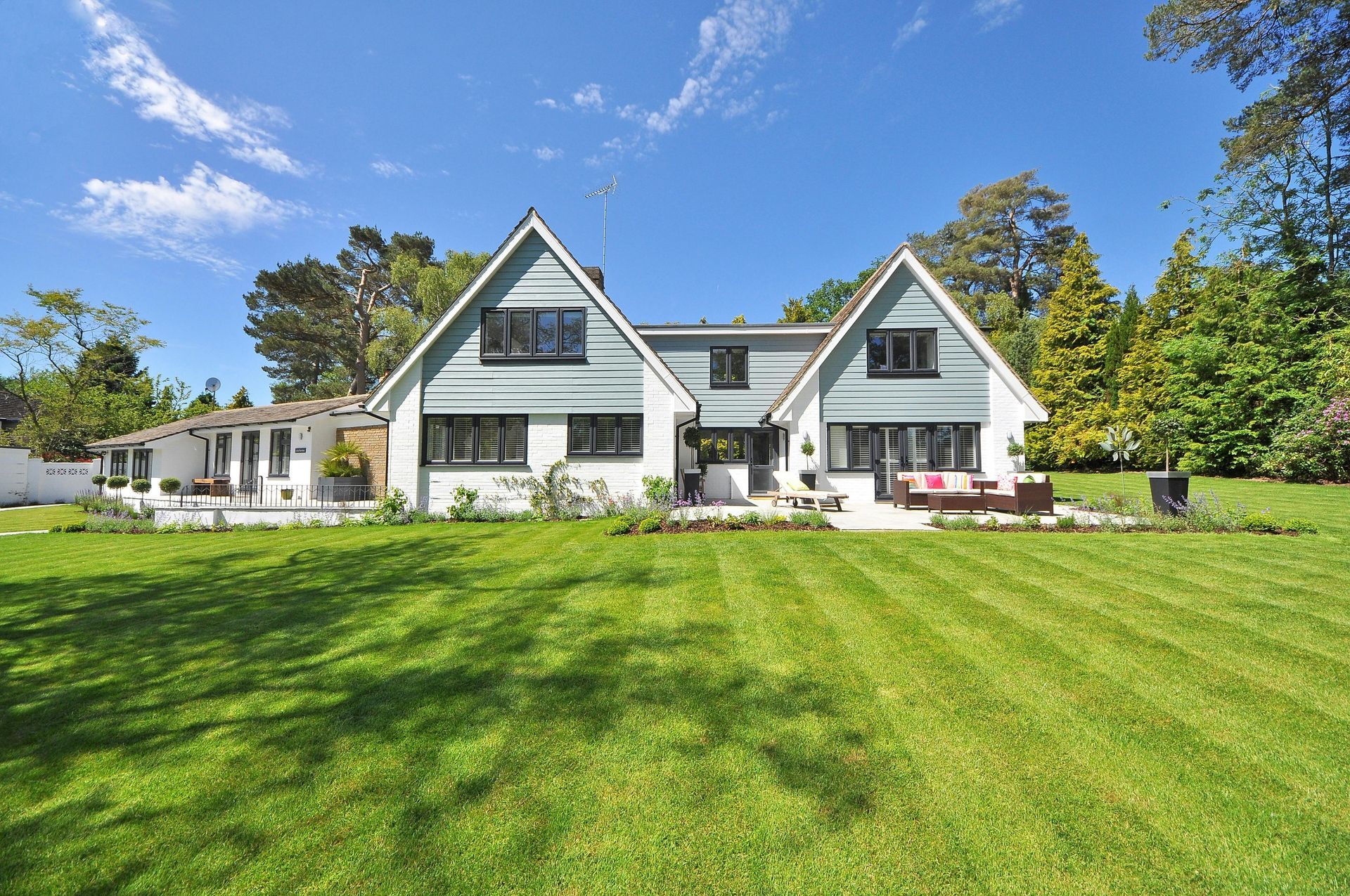 A two-story white house with blue gables, black window frames, and a large manicured lawn under a sunny blue sky.