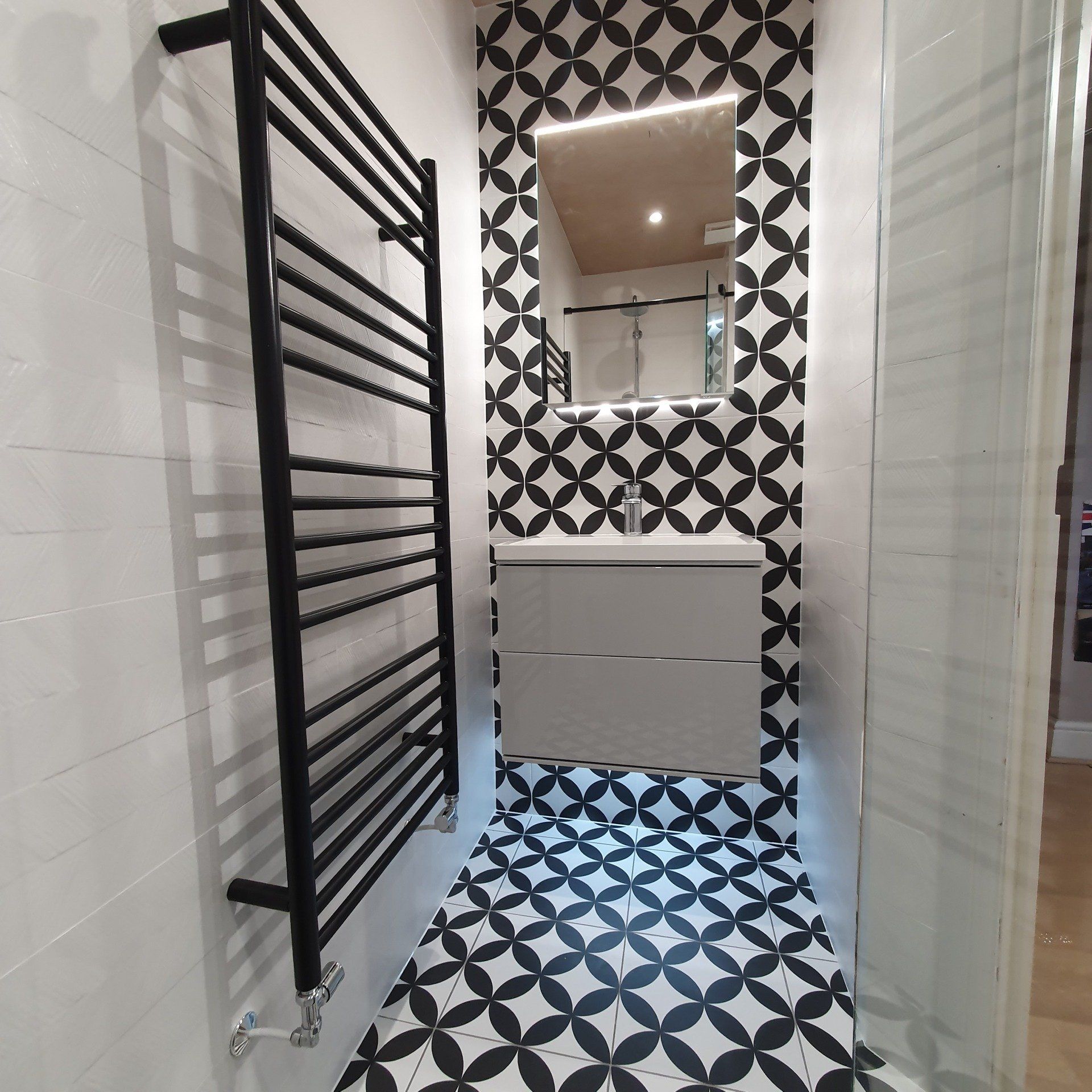 Narrow bathroom with black towel radiator, glass shower and geometric floor tiles.