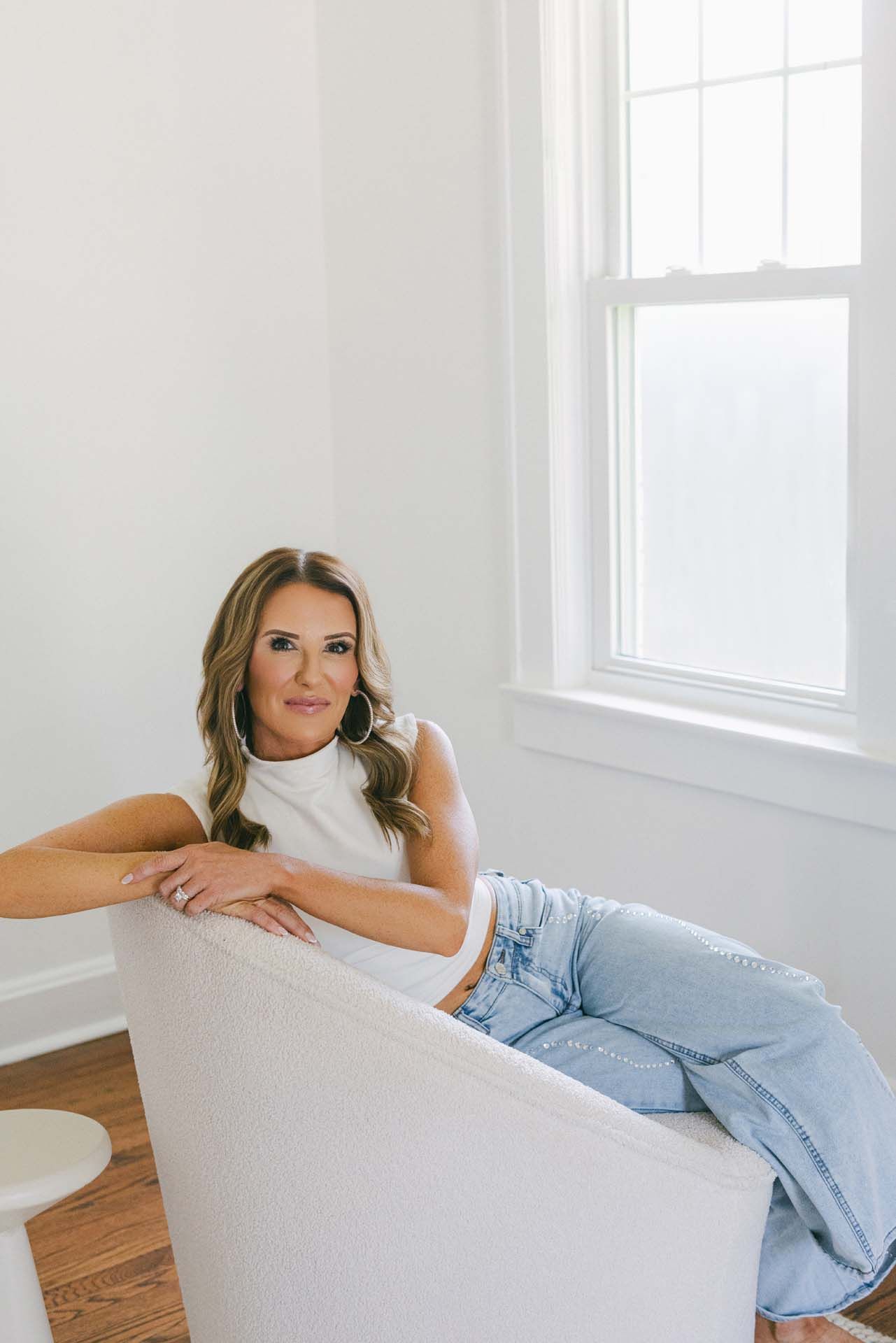 Woman reclining on a white chair in a bright room beside a window, wearing a white top and light jeans.