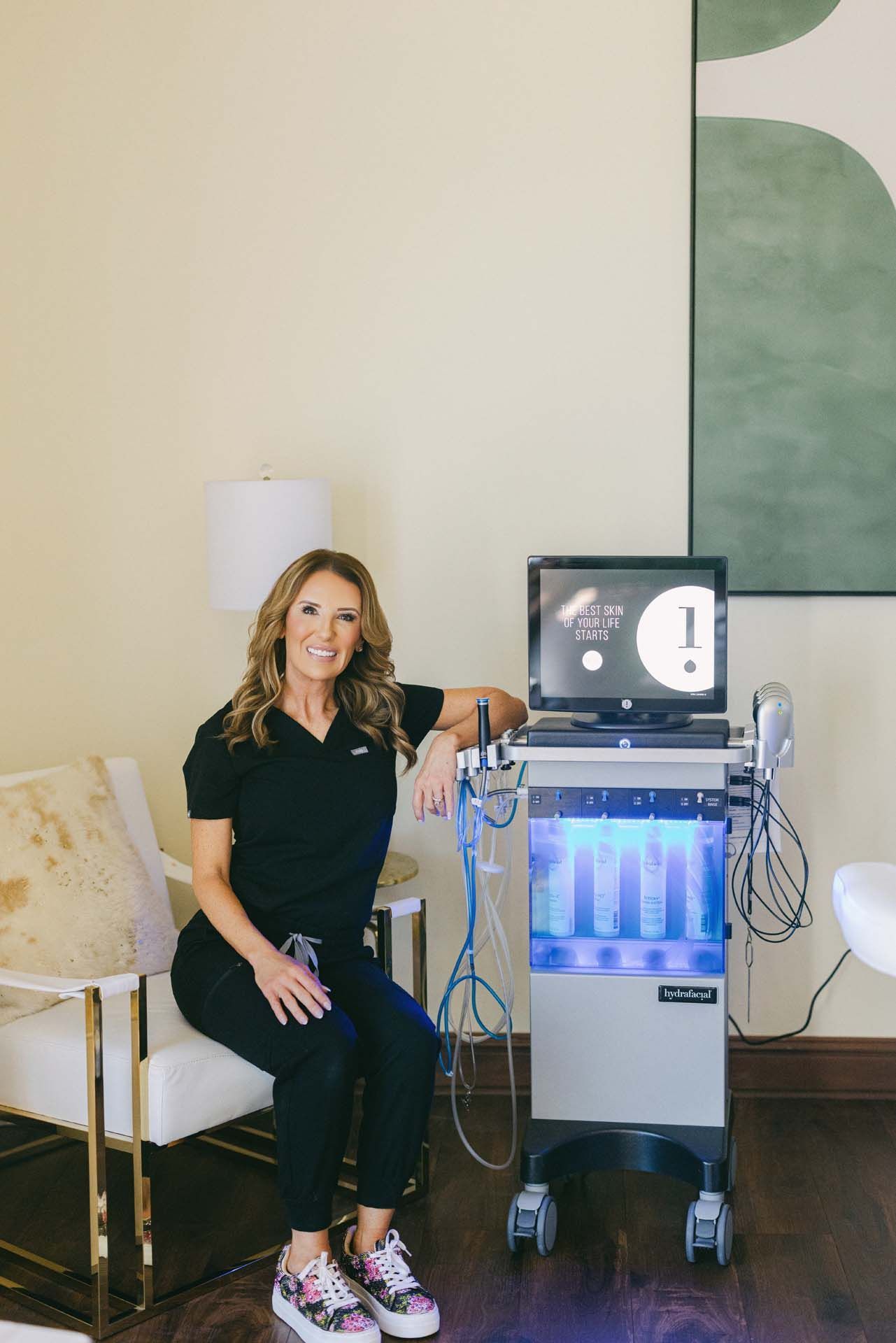 Woman seated beside medical equipment in a clinic room, with monitors and cables visible.