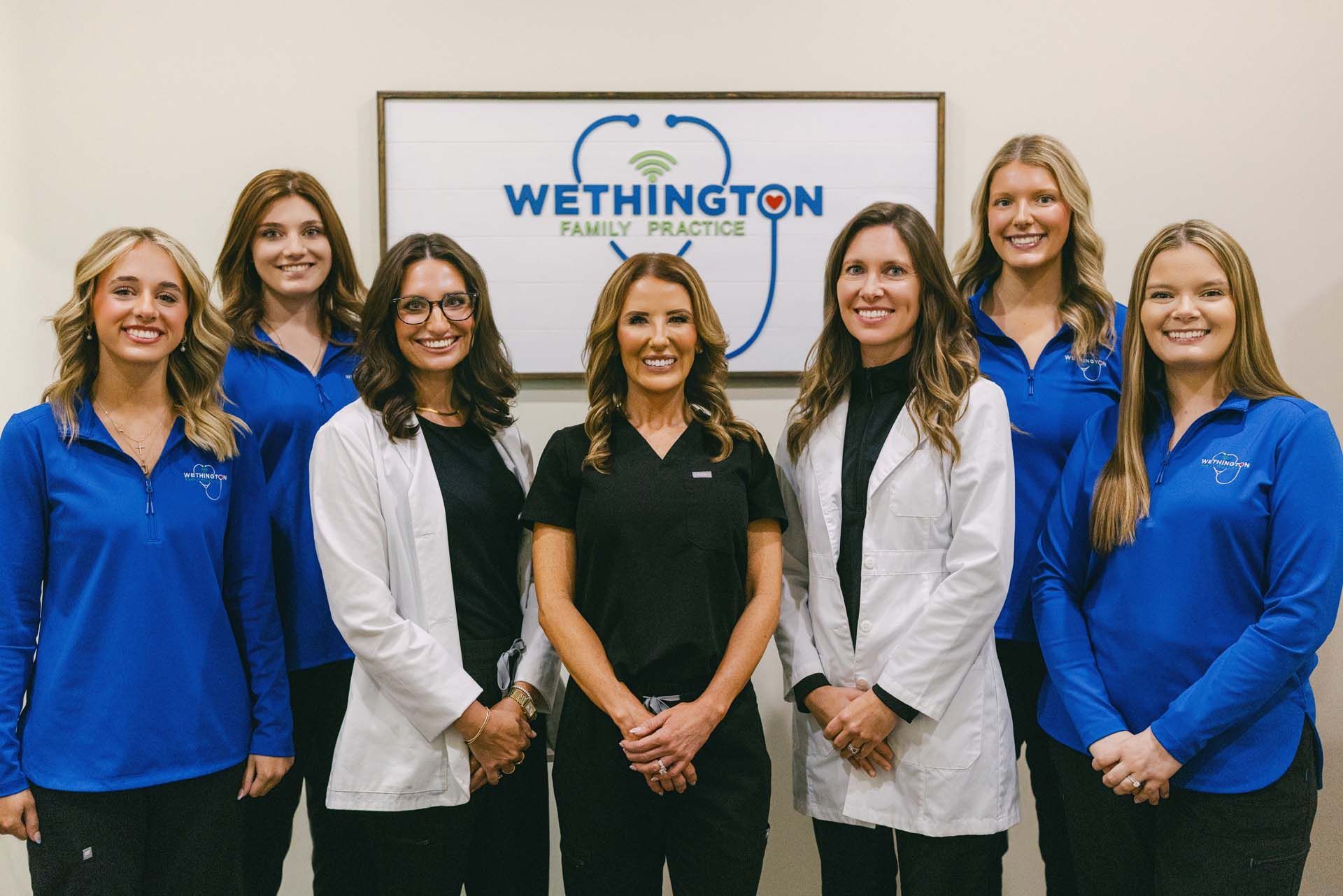 Seven women standing in a row in front of a Wethington Healthcare sign, wearing blue and white uniforms.