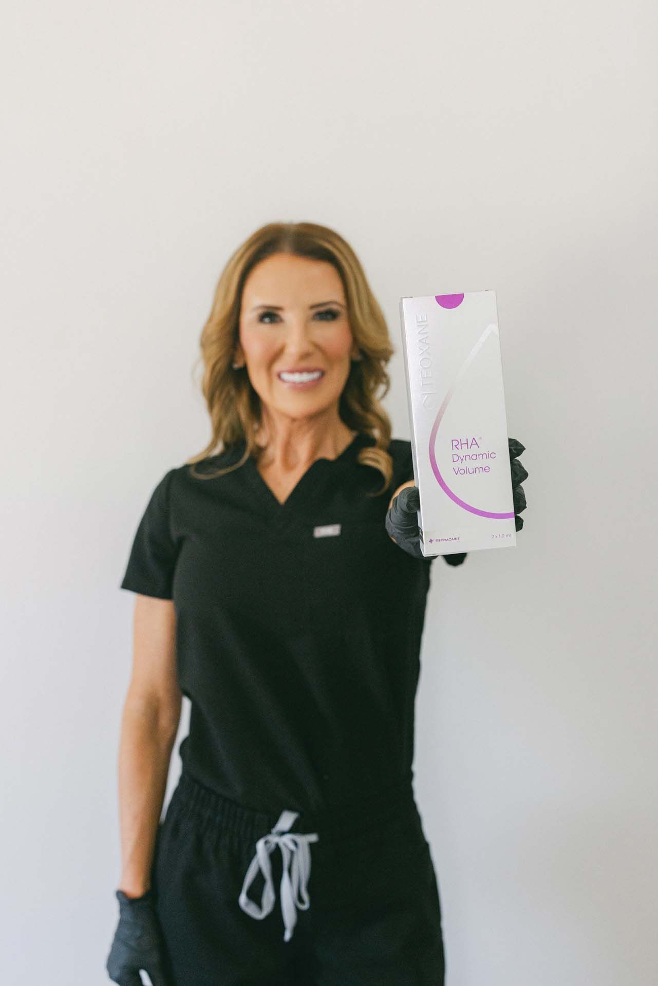 Woman in black scrubs holding up a white product box against a plain wall.