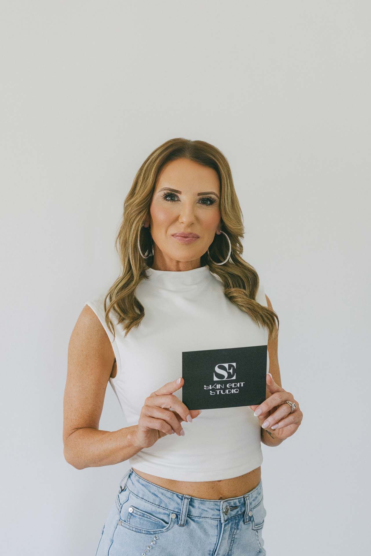 Woman in white top and jeans holding a black card against a plain light background