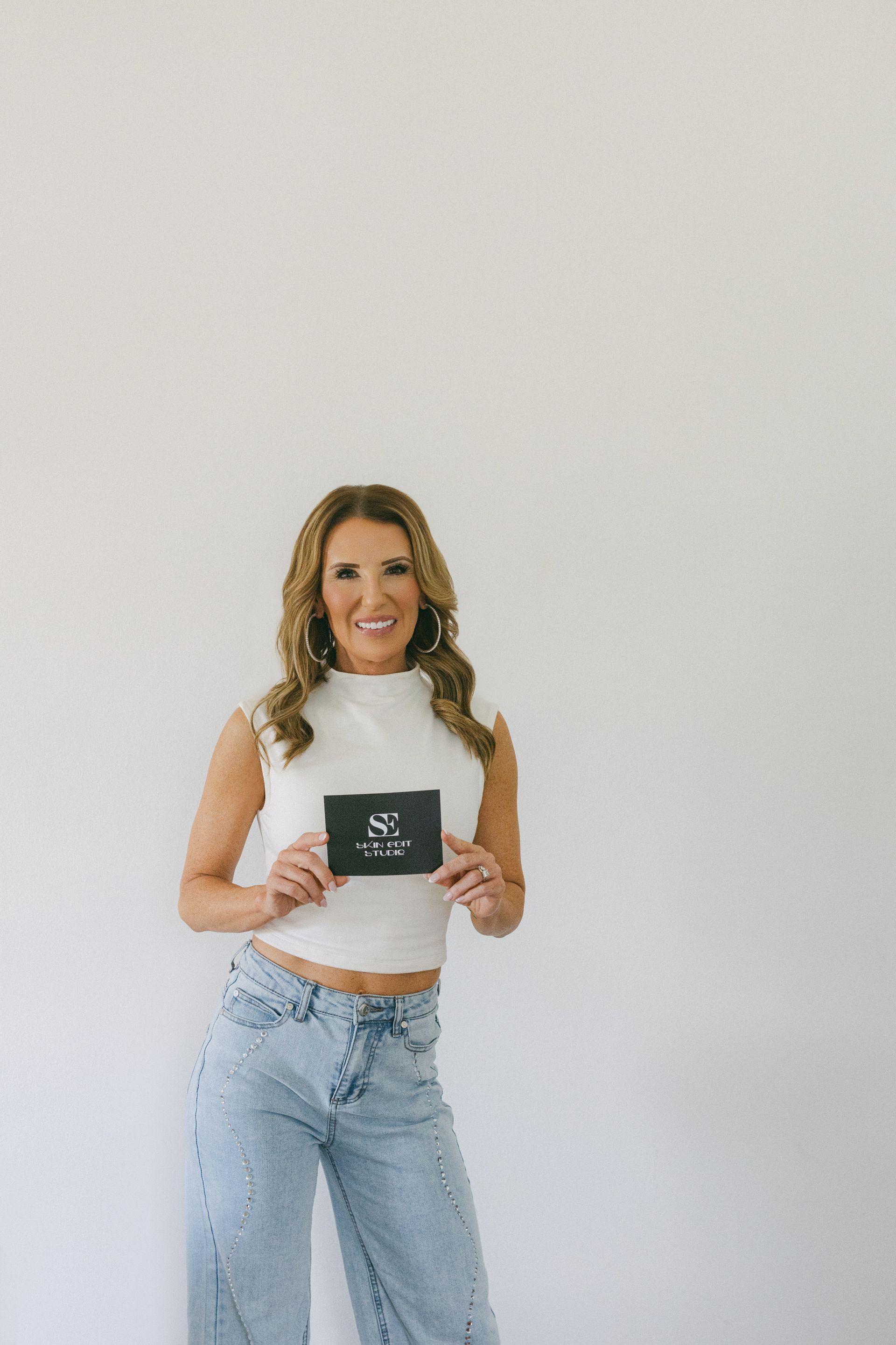 Woman standing against a white wall, holding a black mug and wearing a white top and light blue jeans.