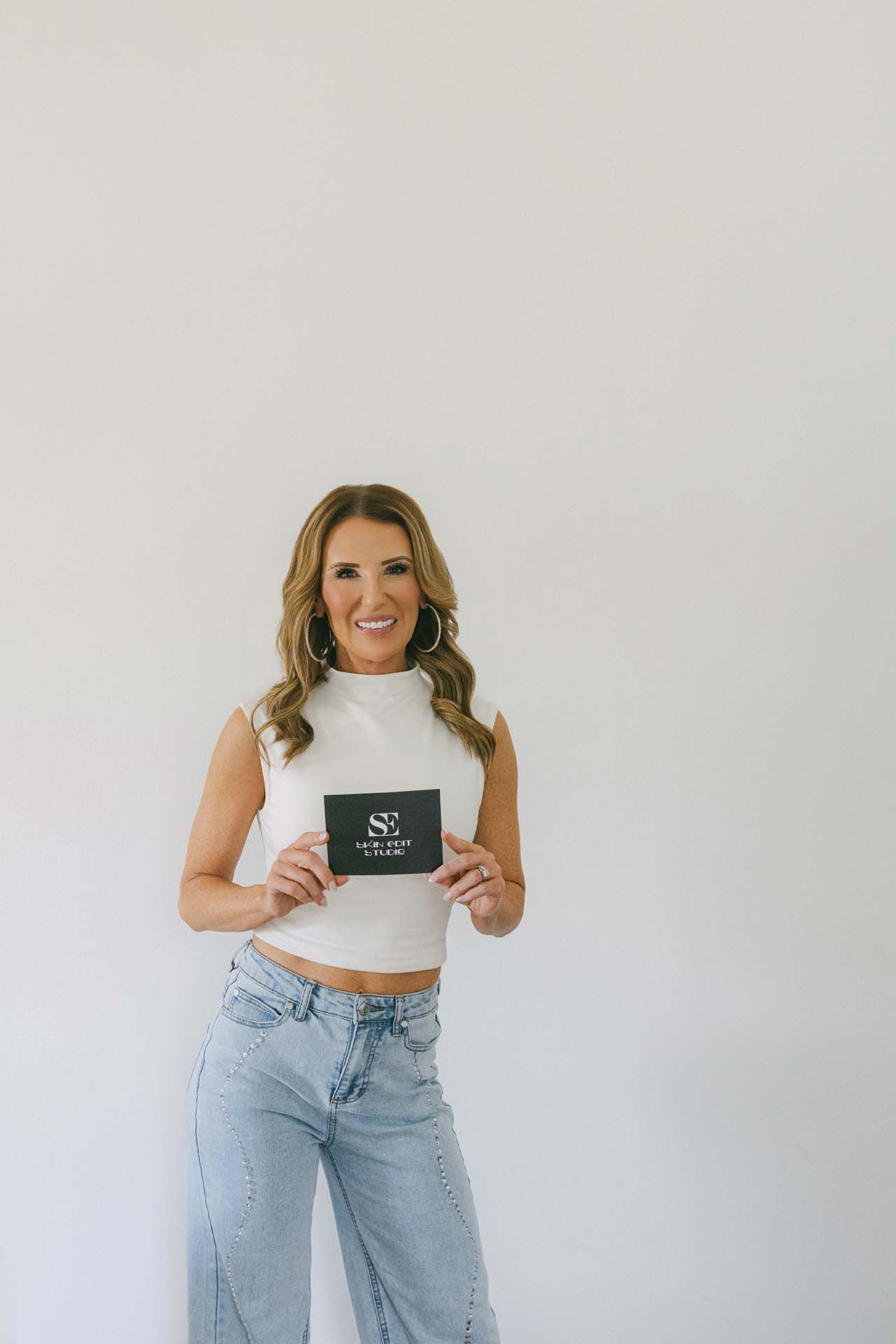 Woman in white sleeveless top and light jeans holding a small black box against a plain white wall.