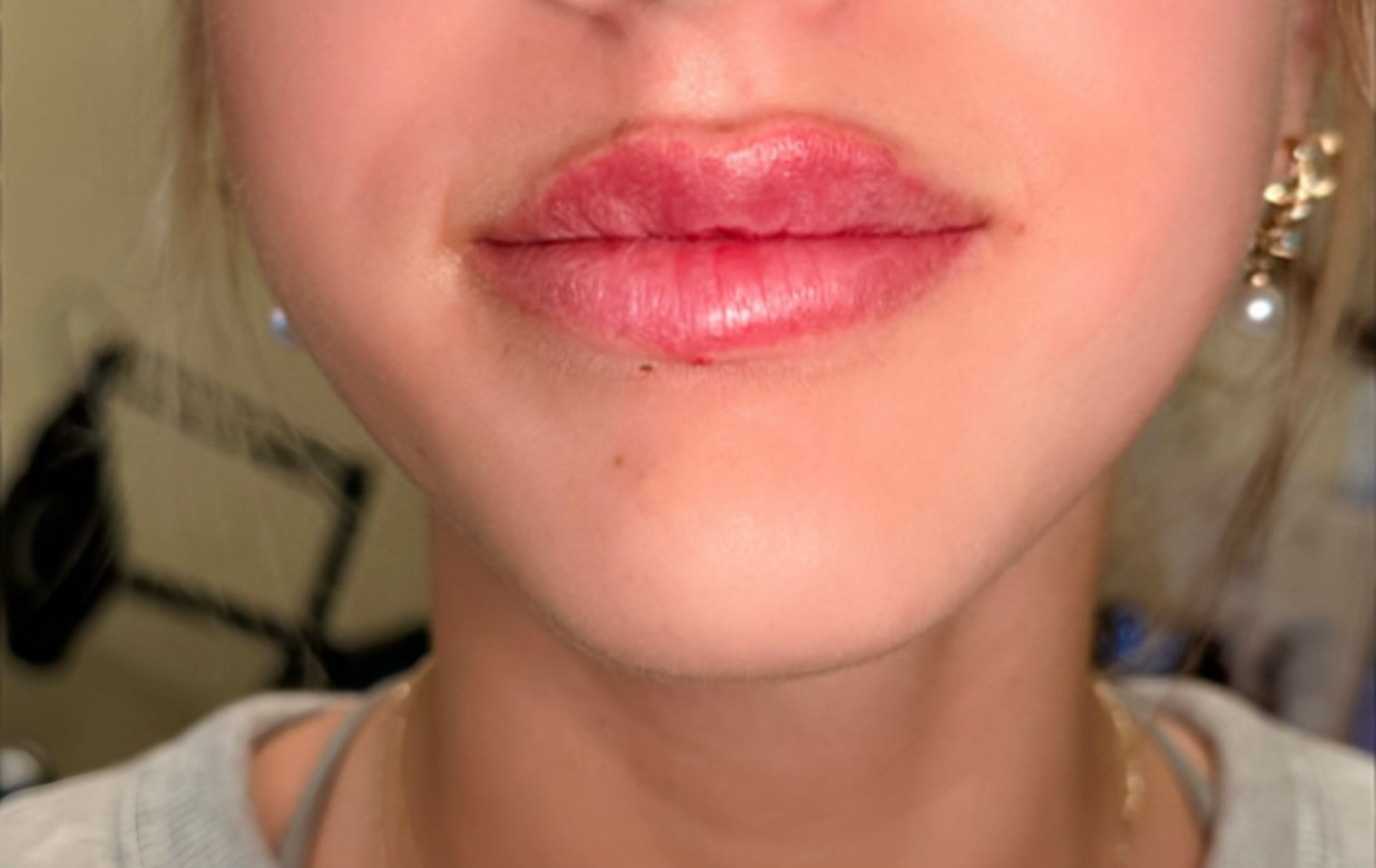 Close-up of glossy pink lips and lower face with gold earrings against a blurred indoor background