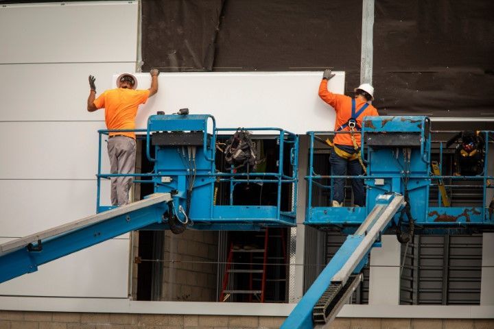 Two workers in orange safety vests stand on a blue aerial lift outside a building facade.