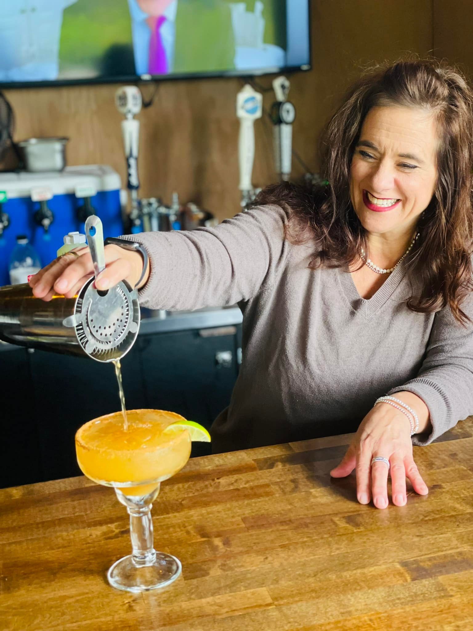 Bartender pouring a drink into a margarita glass. The drink is yellow, behind a bar, smiling.
