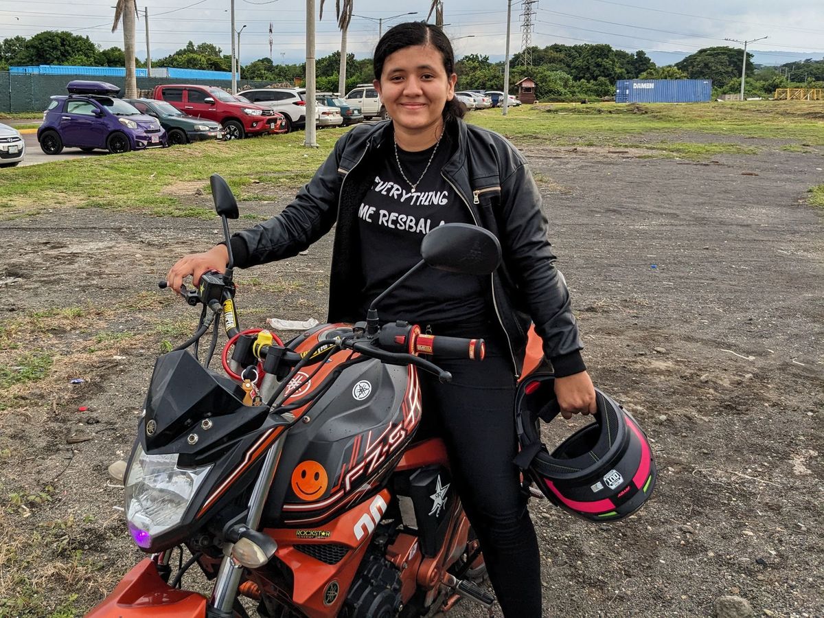 A rider in Nicaragua holding a helmet beside an orange motorcycle with a smiley face sticker on the tank