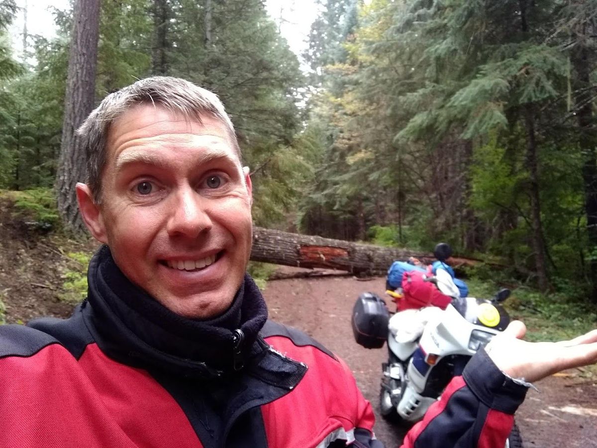Steve shrugging beside a motorcycle with a giant fallen log blocking the forest road