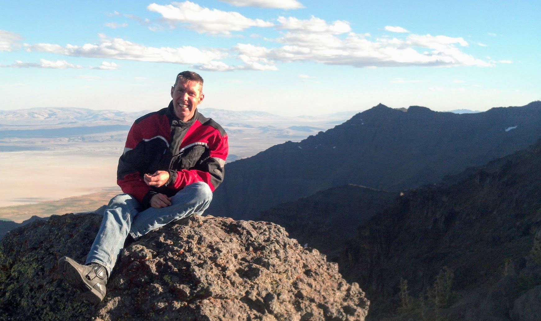 Steve sitting on a rocky mountaintop in a red riding jacket, with a wide valley and dark mountains behind him