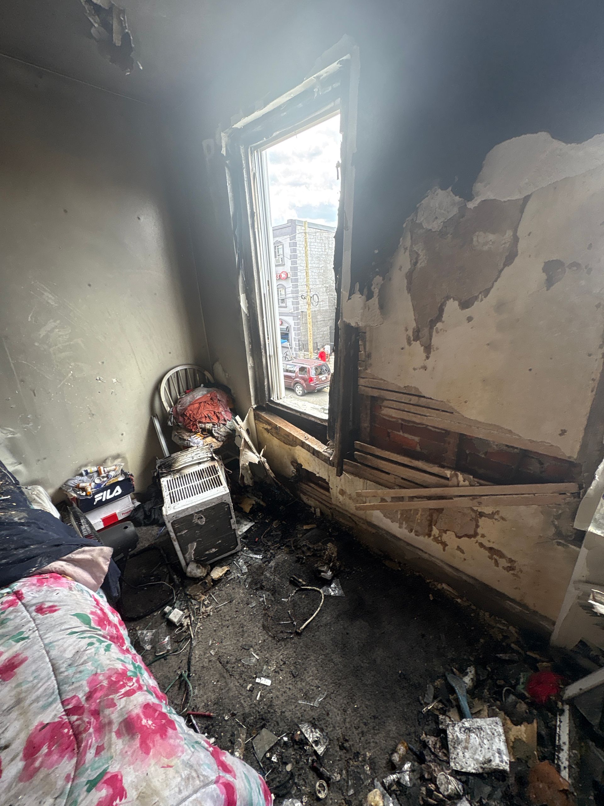 Burned bedroom with blackened walls, charred floor, and damaged window letting in daylight