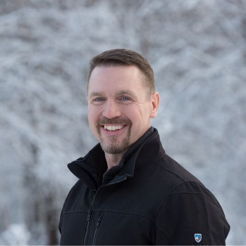 Man smiling, outdoors in winter. Wearing a black jacket, with snow-covered trees in the background.