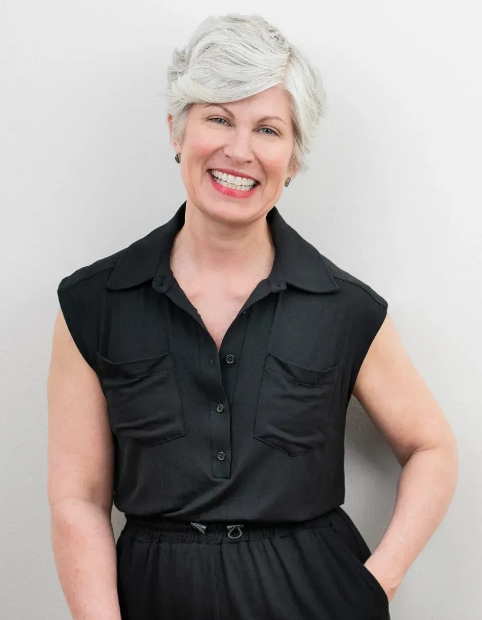 Woman with short, gray hair smiles, wearing a black sleeveless jumpsuit, leaning against a white wall.