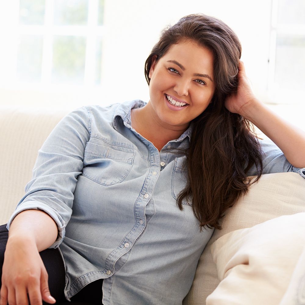 Woman smiling on a white couch, wearing a blue button-up shirt and black pants, hand in hair.