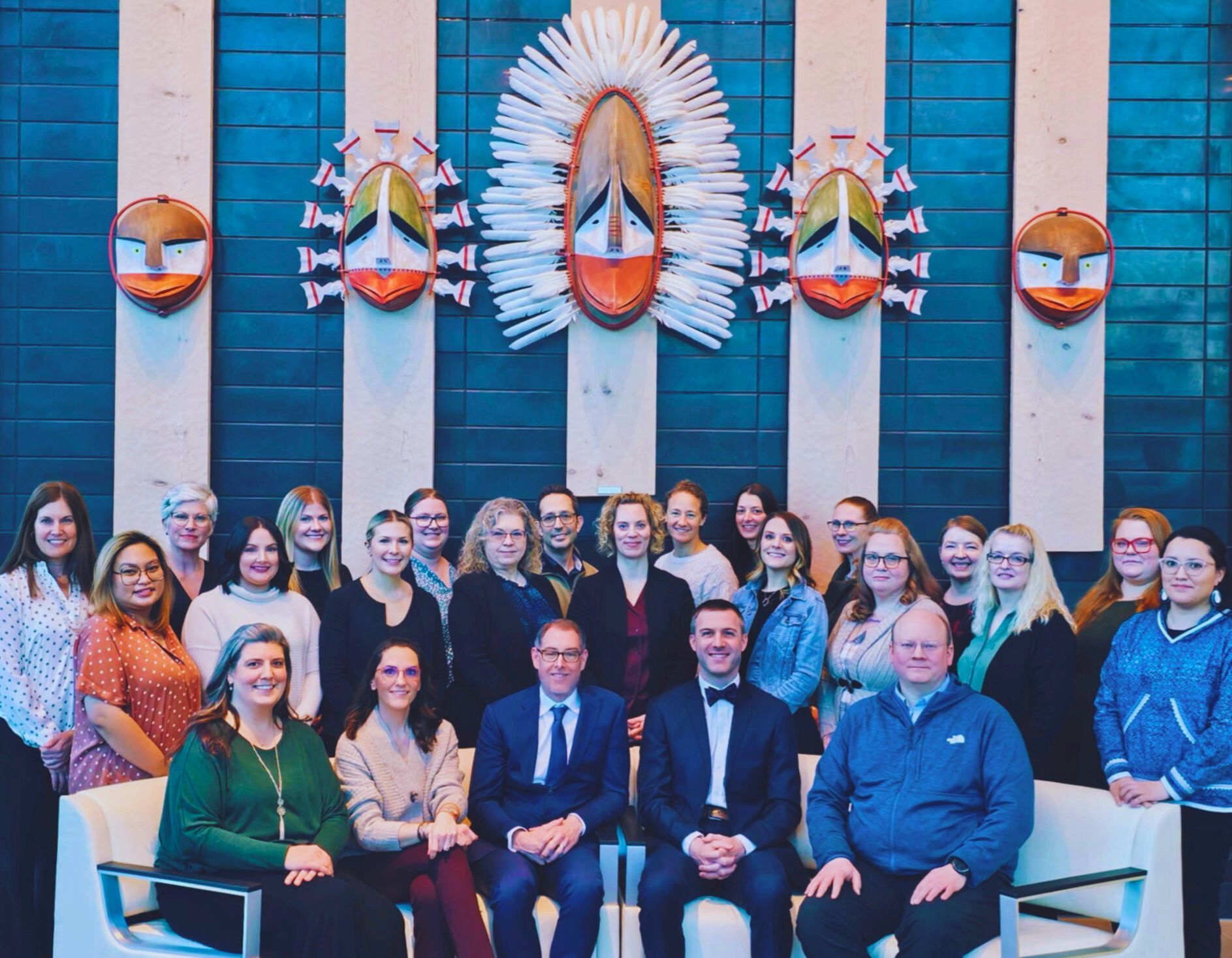 Group photo in front of art. People smiling, some seated. Blue wall with white stripes and art.