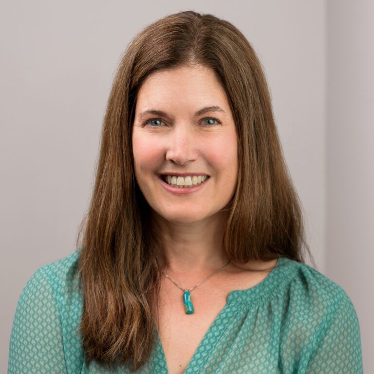 Woman with long brown hair, smiling, wearing a turquoise blouse and necklace.