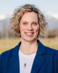 Woman with curly blonde hair smiles, wearing a blue blazer and white shirt. Mountains are in the background.