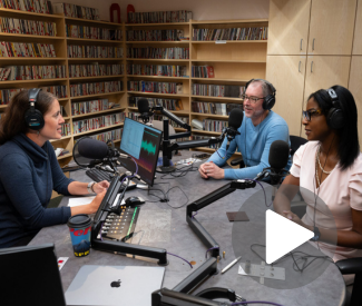 Three people at a podcast recording table in a studio. Microphones and audio equipment are present. Shelves of CDs are visible.