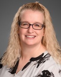 Woman with glasses smiles; blonde hair, patterned top, against gray backdrop.