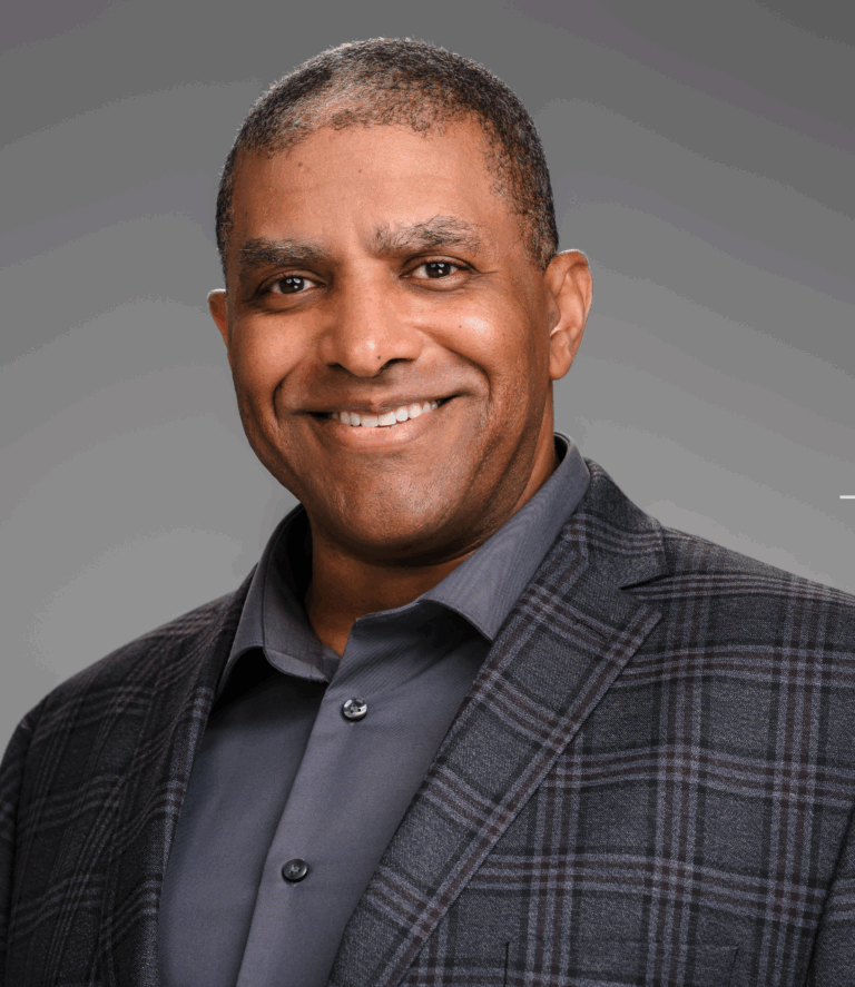 Man in gray shirt and plaid jacket smiles broadly against a gray background.