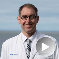 Man in white coat, glasses, smiling; Duke Surgery logo visible, outdoor setting.