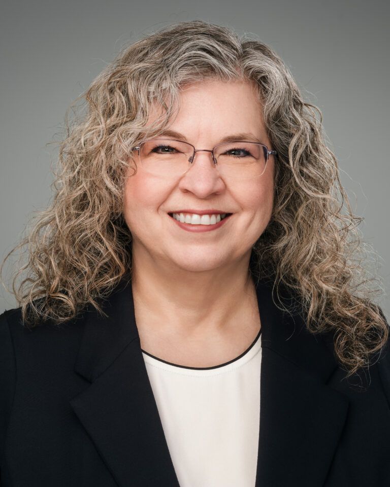 Woman with curly gray hair, wearing glasses, smiling, in a black blazer and white blouse, against a gray background.