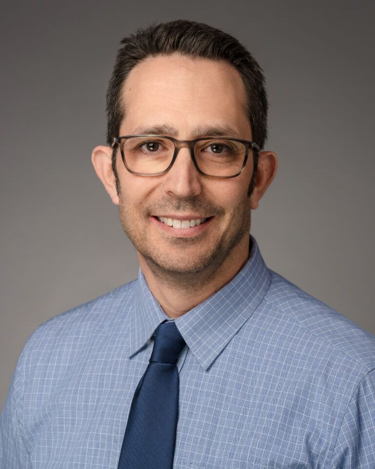 Man in glasses and blue shirt, smiling, headshot.