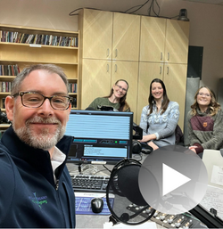 Man in glasses smiles at the camera in a radio studio with 3 women smiling in the background.
