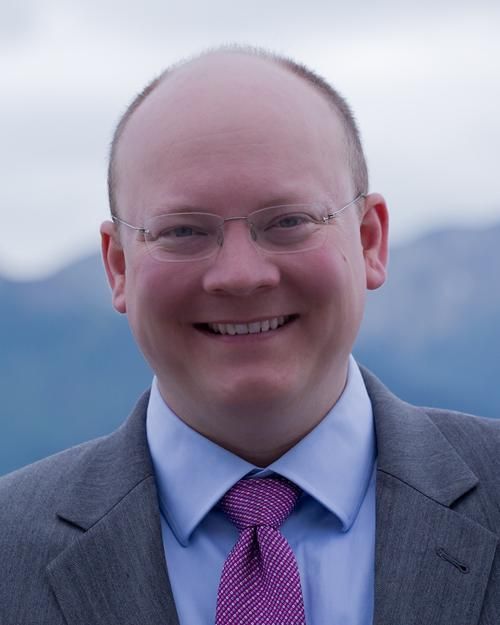 Man in a suit smiles, wearing glasses and a purple tie, with a mountain backdrop.