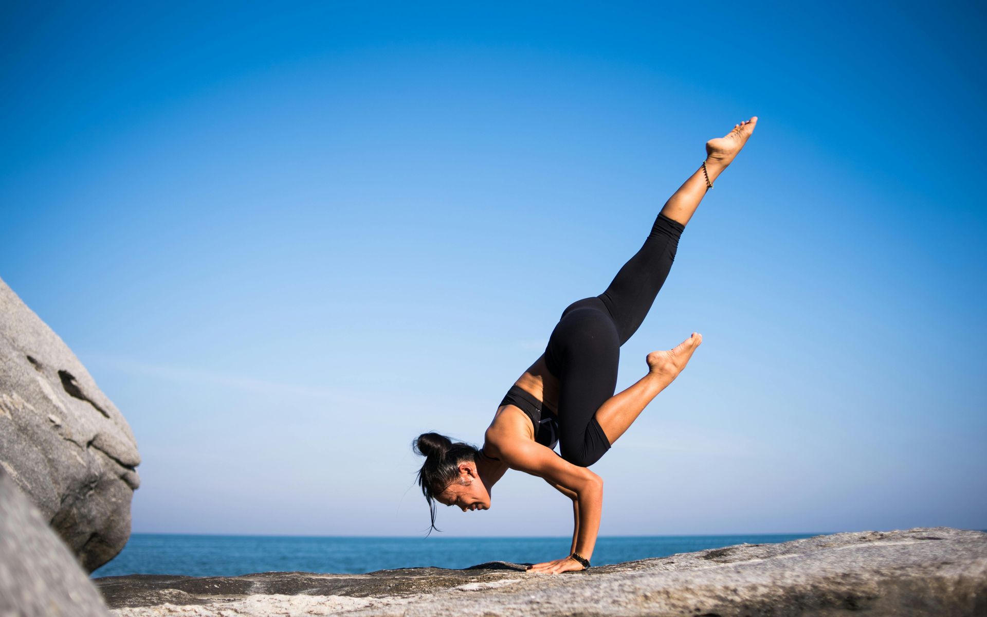 Woman doing a yoga handstand on a rock near the ocean; blue sky.