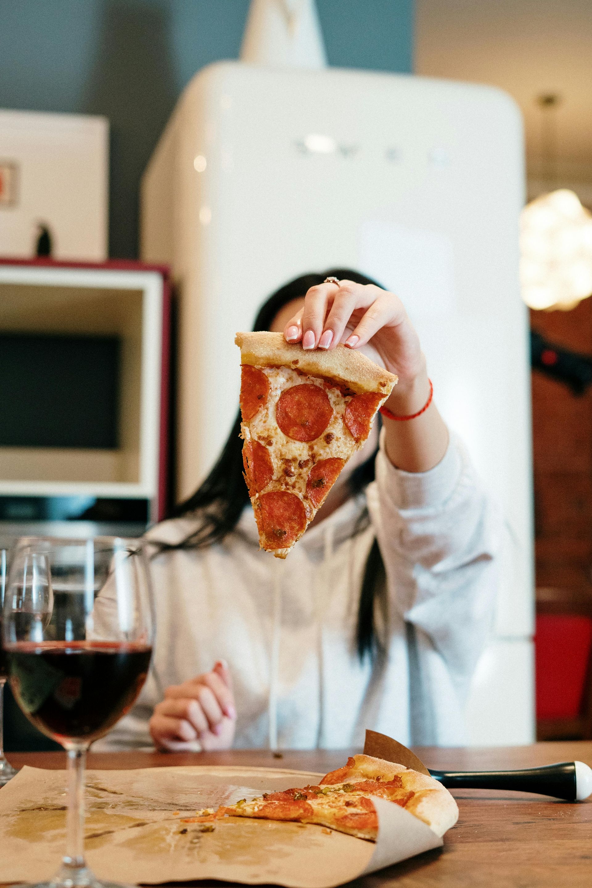 Woman holding up pepperoni pizza slice, red wine glass in front, near a retro refrigerator.