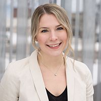 Woman with blonde hair, wearing a cream blazer and a black top, smiling, necklace, in a modern office setting.