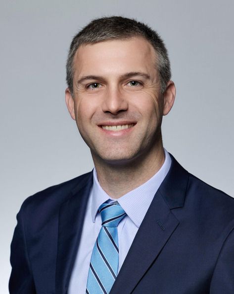 Man in navy suit and blue striped tie smiles, shoulders up, against a gray backdrop.