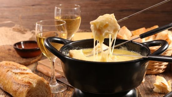 Fonduta di formaggio in una pentola nera con pane, cubetti di formaggio e due bicchieri di vino bianco su un tavolo rustico.