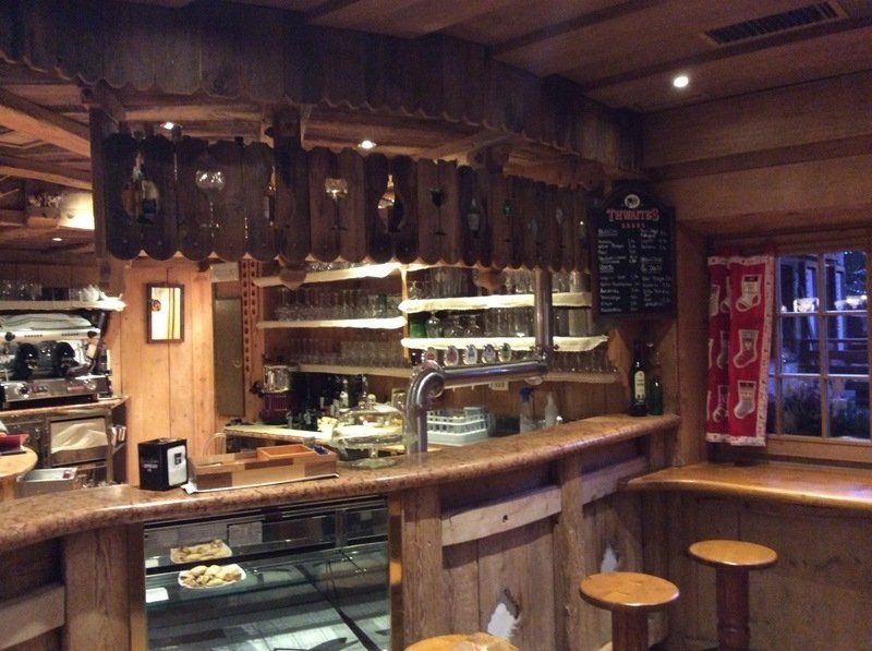 Accogliente interno da bar in legno con sgabelli, scaffali per bottiglie e un bancone espositivo in vetro.