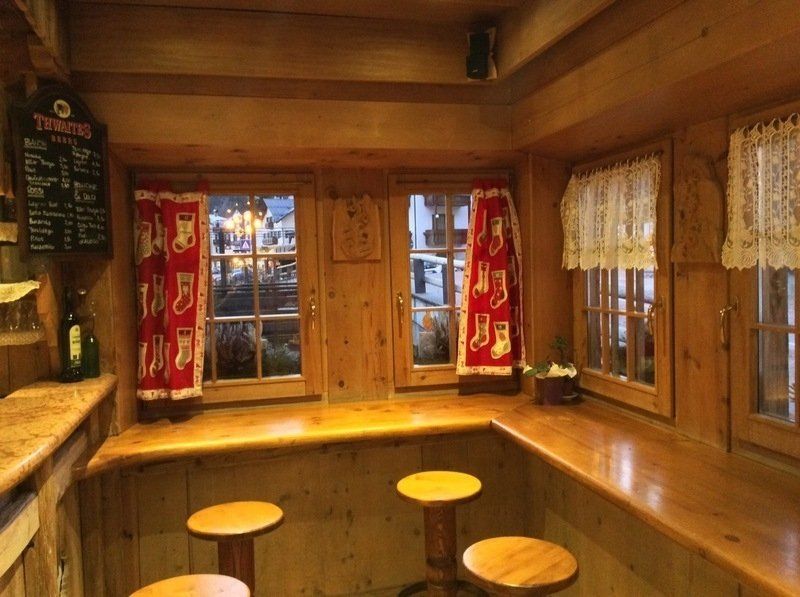 Accogliente interno di una baita in legno con panche, sgabelli, tende rosse e finestre.