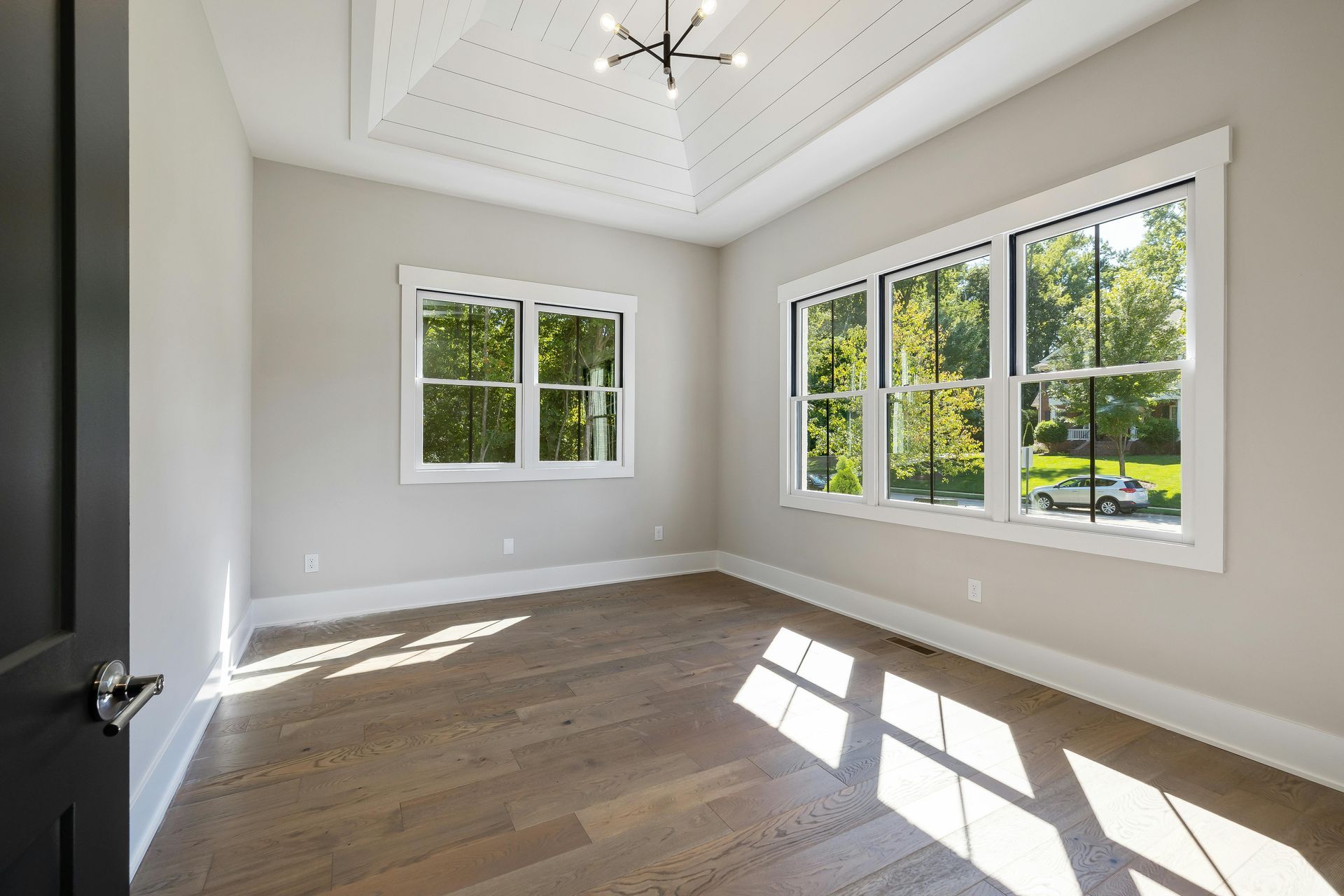 Empty room with light wood floors, beige walls, windows with white trim, and a tray ceiling with a modern chandelier.