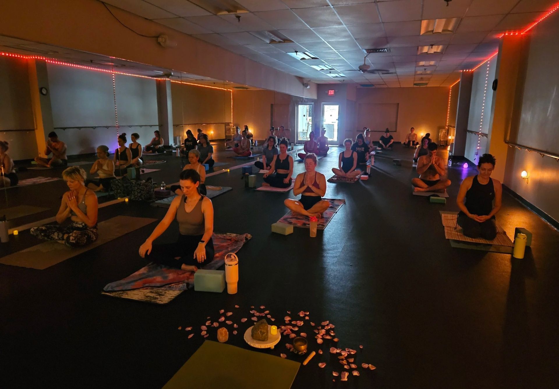 A yoga class in a dimly lit studio with red ambient lighting, people sitting on mats in a circle during a guided practice.