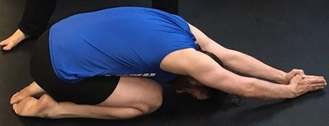 A person in a blue shirt kneels on a dark floor in a child’s pose, with arms extended forward and hands pressed together.