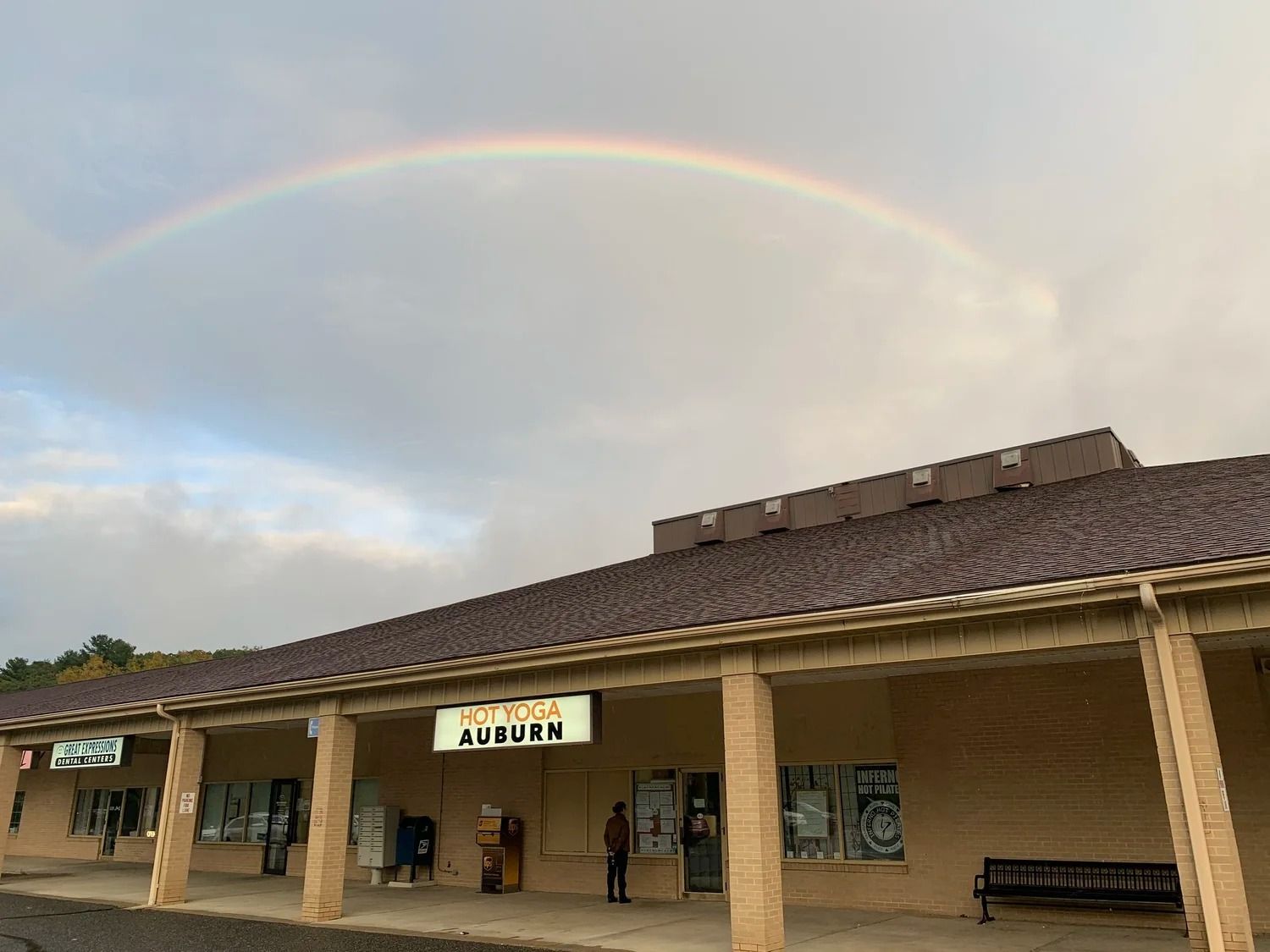 A faint rainbow arches over a tan, one-story commercial strip mall building under a cloudy sky.