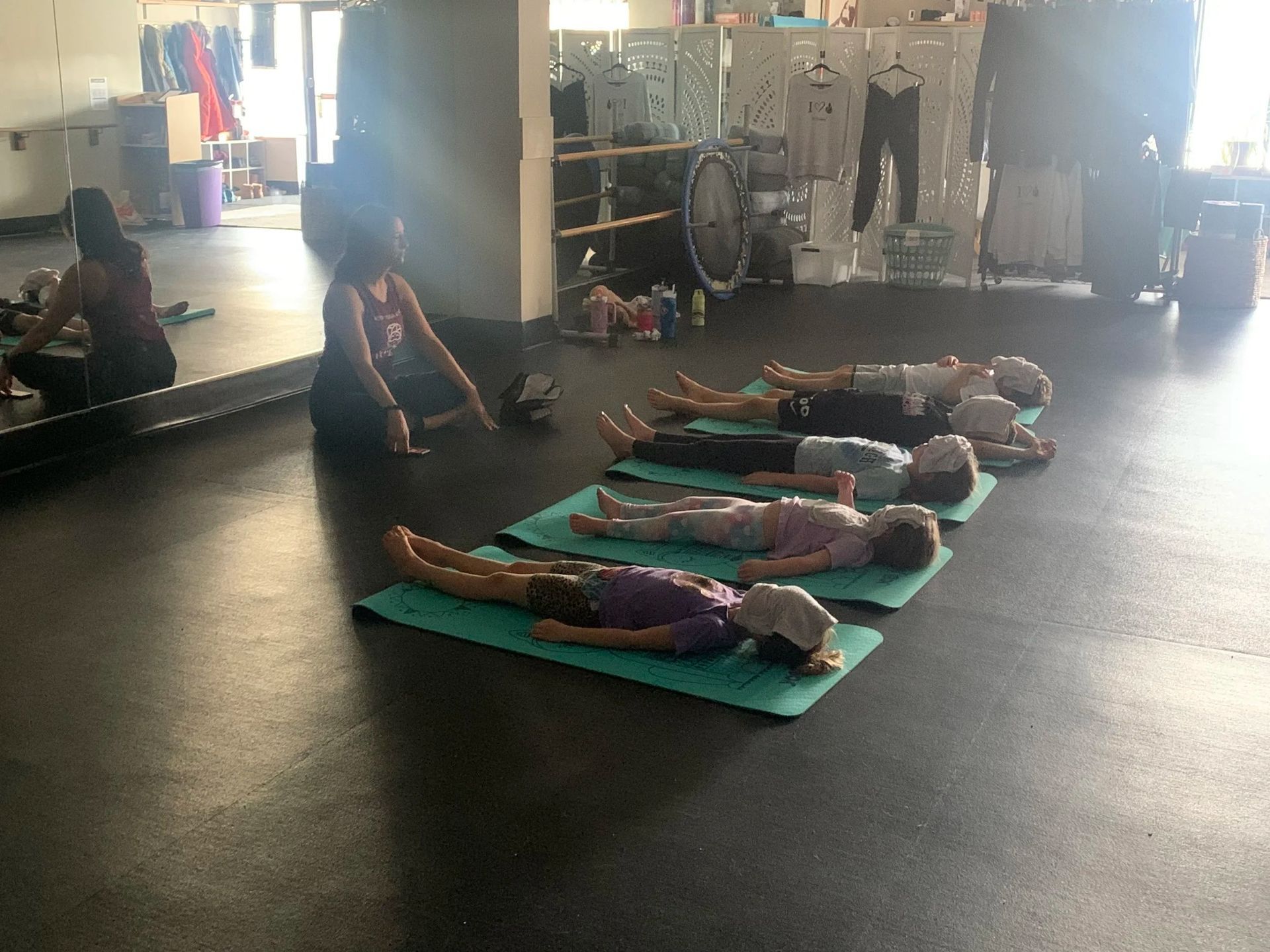A yoga instructor sits in front of a group of people lying on mats in a studio setting.