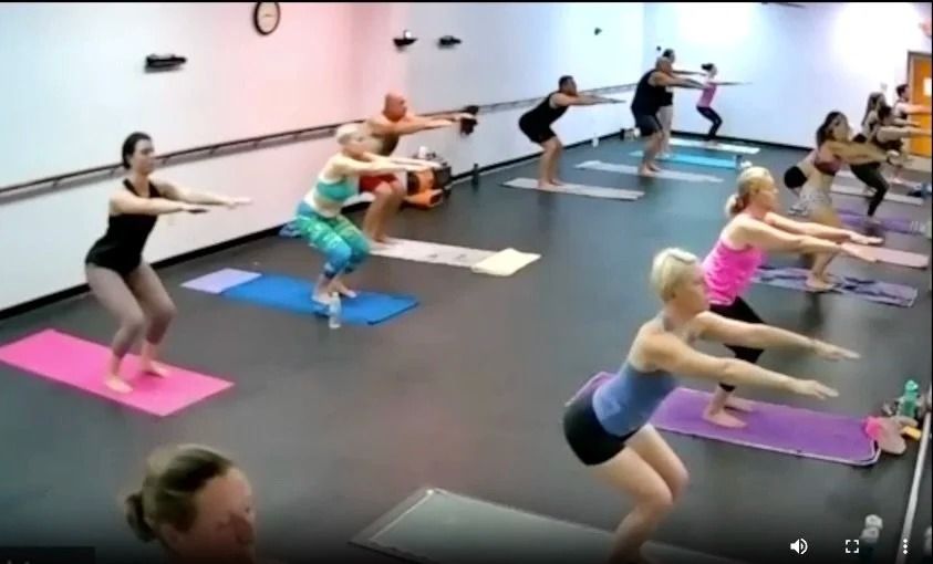 A group of people in an exercise class perform squats on colorful mats in a studio with a ballet barre along the wall.