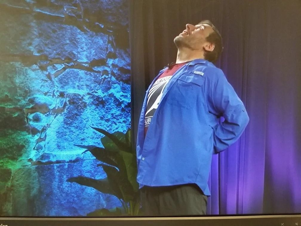 A person in a blue button-down shirt performs a standing backbend stretch with hands on their lower back.