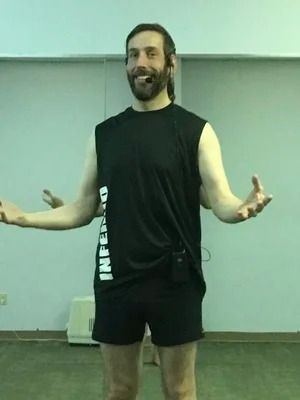 A smiling person in a black tank top and shorts stands in a room, arms extended with palms open.